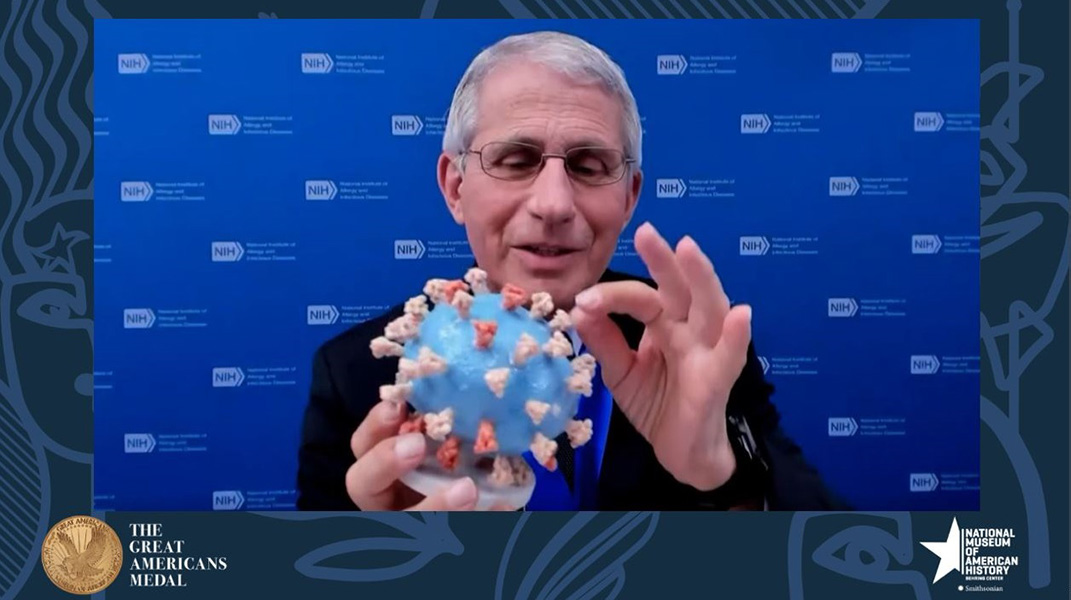 Photograph of Anthony Fauci holding an educational coronavirus model: It is a round blue blob with red spikes protruding from it.