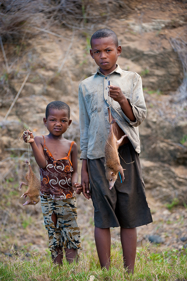 Photograph of two boys standing in the grass, with rocks and dirt behind them. Each is holding a brown, long-snouted dead mammal attached to a piece of twine.