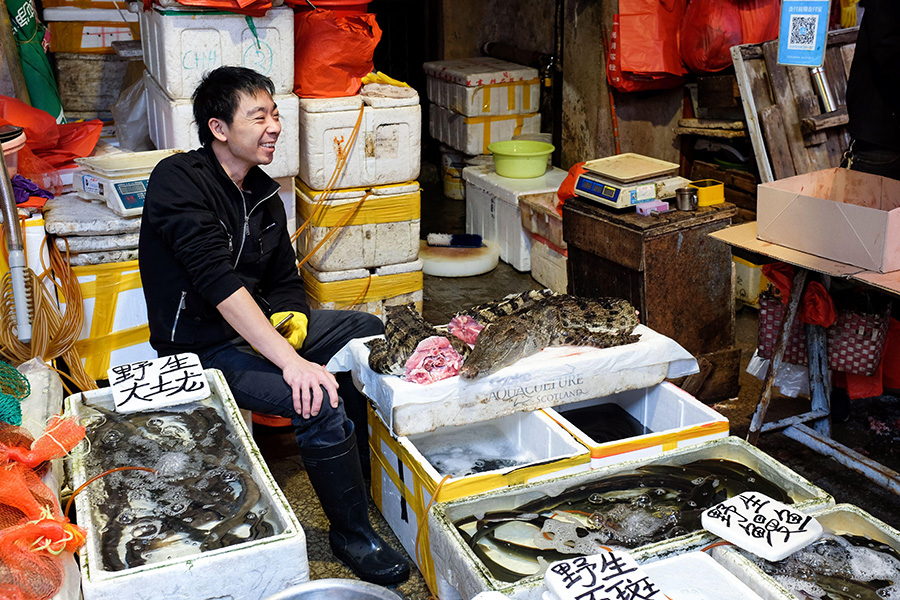 A man in black clothing sits smiling, surrounded by containers and displays of different types of raw flesh. 