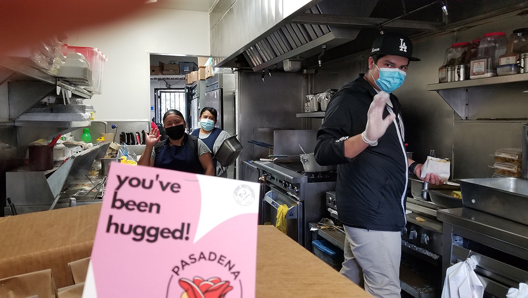 In a restaurant kitchen, three masked workers pause from meal preparation to wave at the camera. A large sign that says, “You’ve been hugged!” is visible in the foreground.