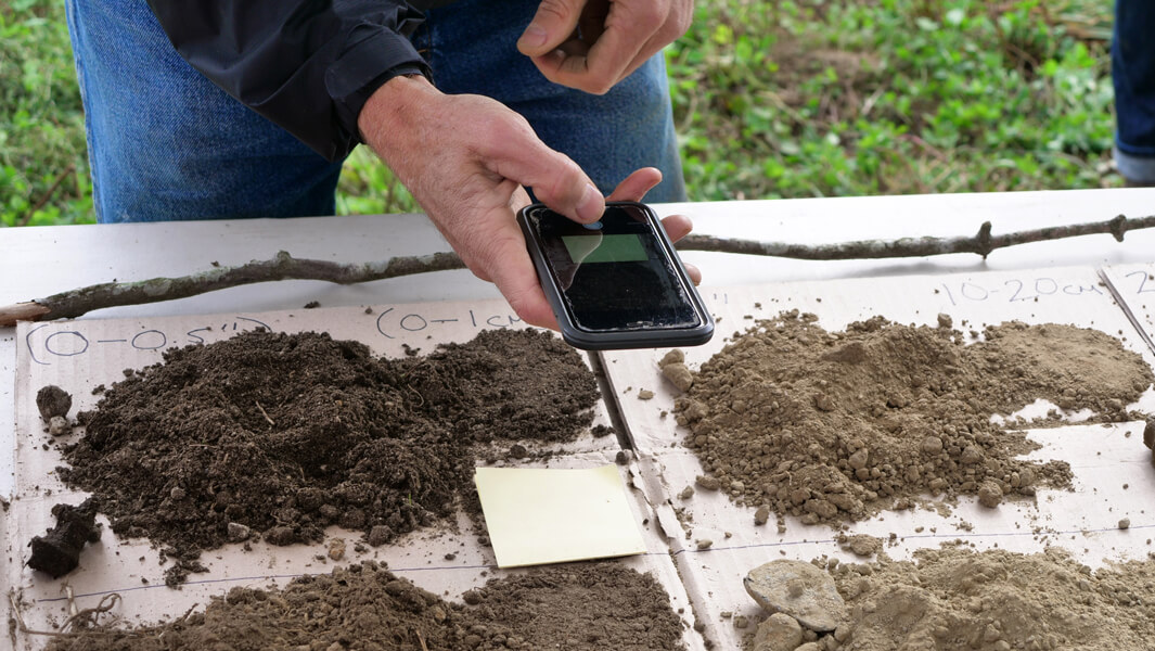 Photograph of a table with four piles of differently colored earth on them. A person’s hand is seen holding a mobile phone over the soil, taking readings.