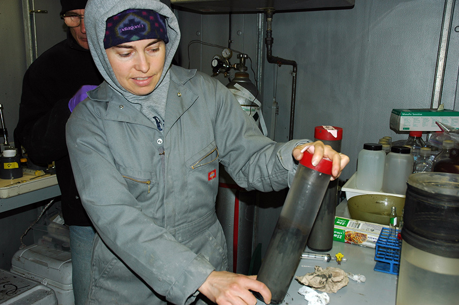 Photograph of a woman in grey sweatshirt and zipped-up jacket. She is holding a cylinder filled with dark, murky fluid. She is in a dark room with grey metal walls; a bench in front of her holds racks, plastic bottles and screwed-up paper towels. A gas tank and another person are in the background.