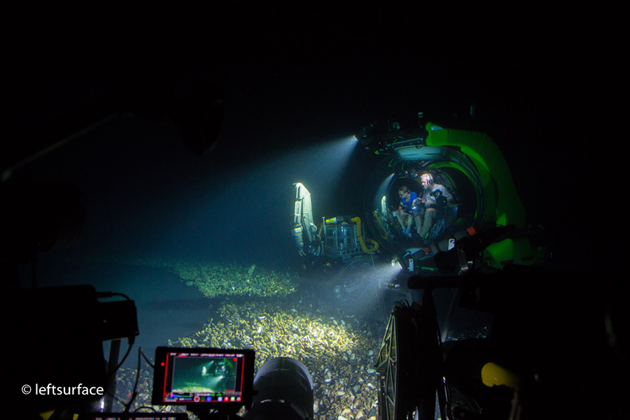 A dark, blurry photograph showing a submersible. Part of the seafloor is lit up by lamps and there are two scientists visible. In the foreground, a person sits in front of a computer monitor; video equipment, deeply shadowed, is in the foreground too.