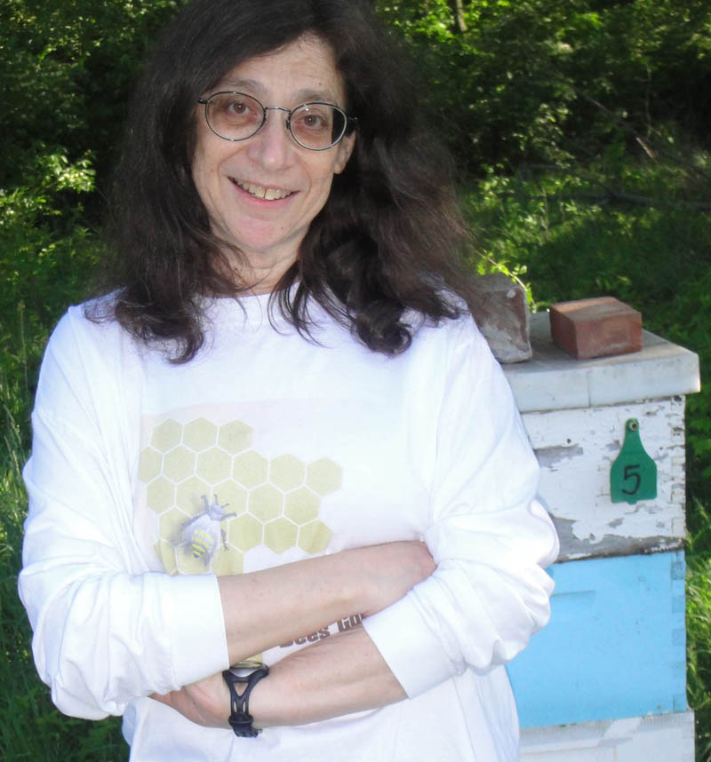 Photo of May Berenbaum wearing white sweatshirt with a honeycomb on it, standing in front of an apiary.