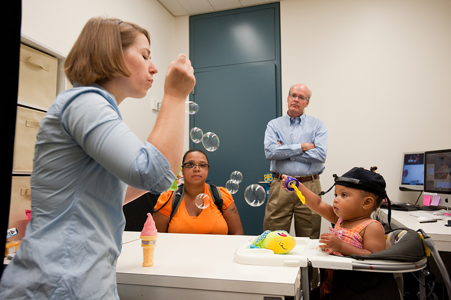 A researcher blows bubbles as a young child and her parent look on