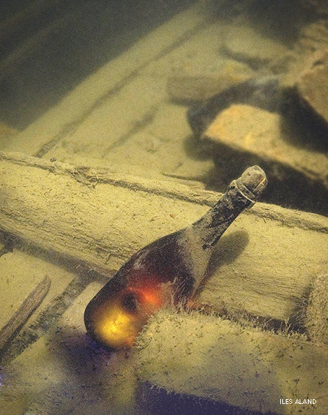 Photograph of a bottle of champagne covered with silt, under water. It is lying in the remains of a ship.