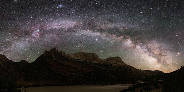 Photograph at night, with mountainous terrain and desert floor. In the sky one can see thousands of stars and the disk of the Milky Way galaxy.