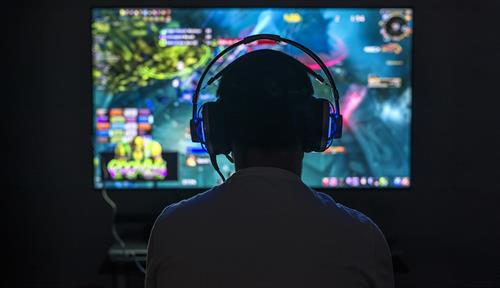 Silhouette of video gamer seen from behind, wearing headset. In front of the gamer is a brightly colored computer screen.
