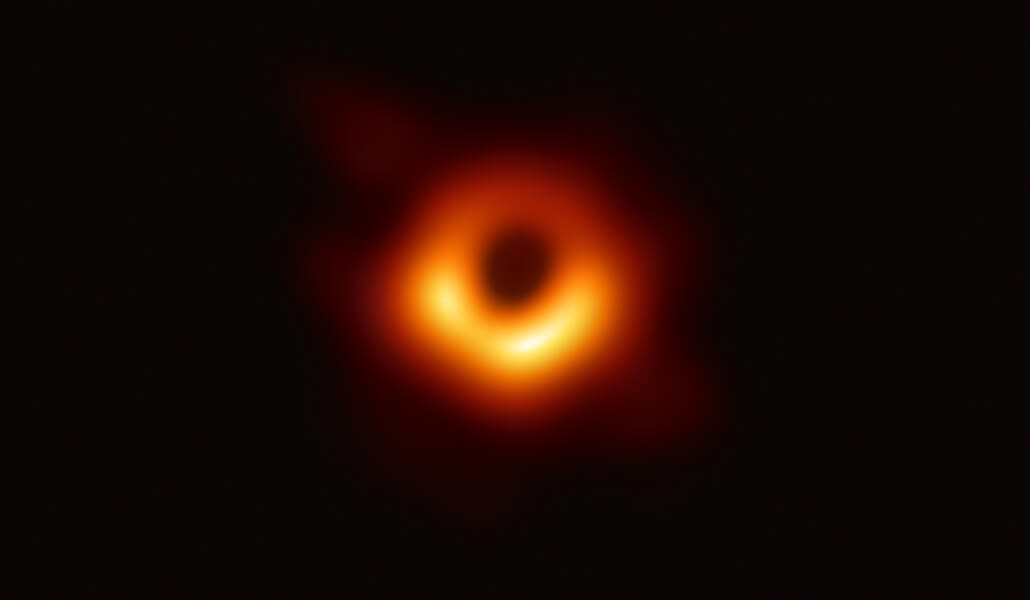 Image shows a dark shadow ringed in bright red and yellow formed as light bends in the intense gravity around a black hole.