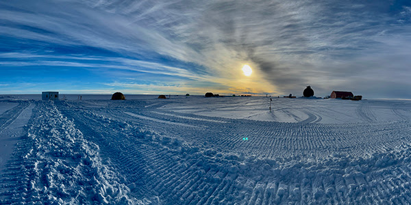 Photograph of a flat, desolate, ice- and snow-covered landscape with the Sun low in the sky. A few structures are in the background.