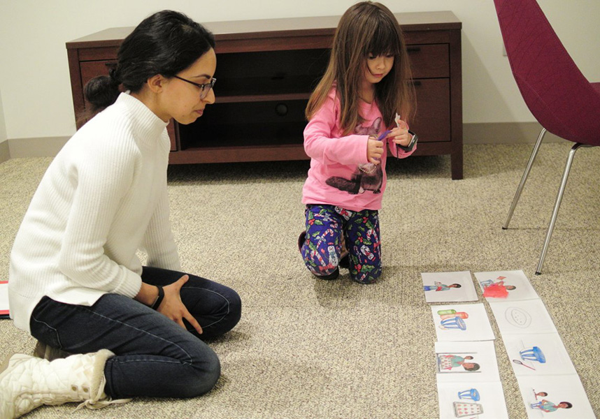 In a test of recall, an adult researcher looks on as a young girl look studies a series of pictures from a story book the girl had listened to earlier.