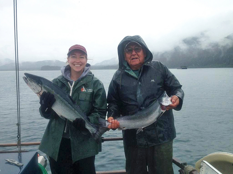 A woman and a man pose holding each a salmon they have caught. In the background you see water and a mountain covered by clouds.
