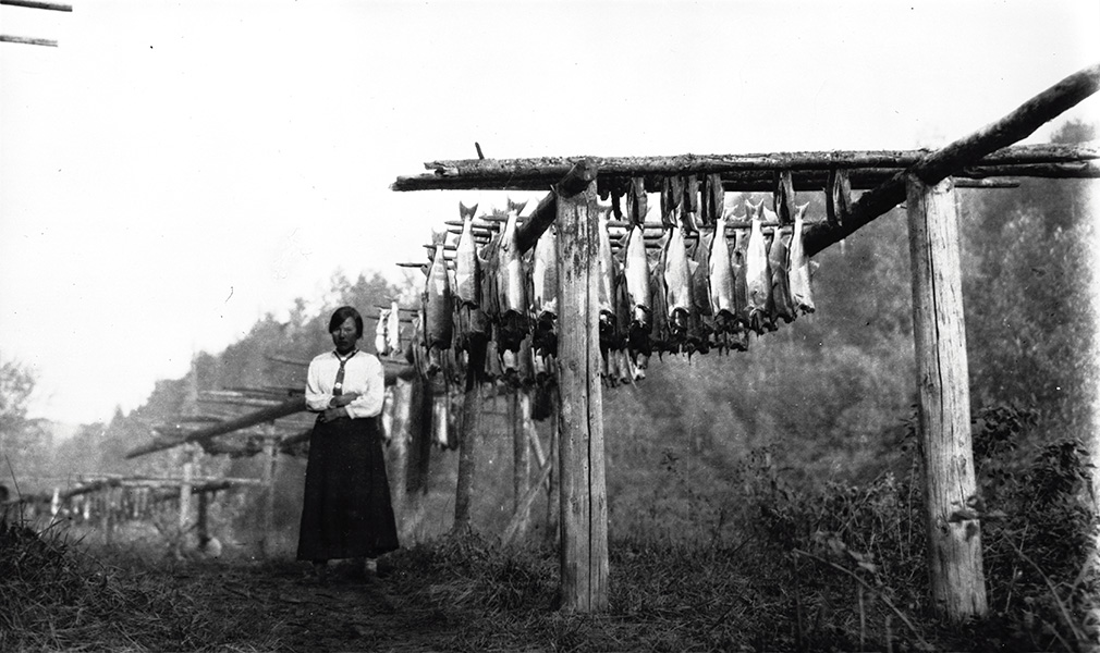 A black and white image captures a woman standing next to a salmon drying rack with dozens of fish.