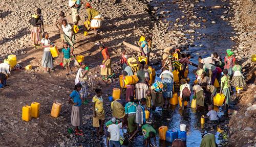 A group of about three dozen people stand in and around a river near Konso, Ethiopia, collecting water in large blue or yellow plastic containers.