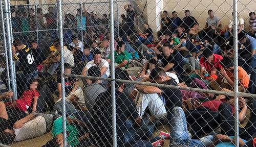 Photo shows many people being held within a chain-link fence inside a building.