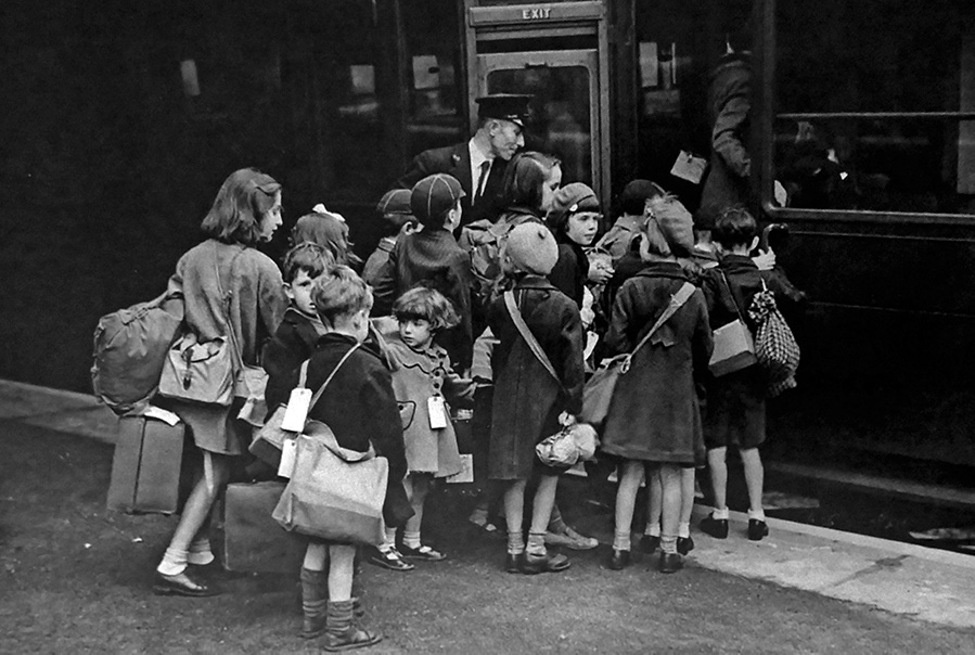 Black and white historical photograph of a crowd of about a dozen young children, each wearing an identity tag, boarding a train. Satchels and bags are slung over their shoulders.