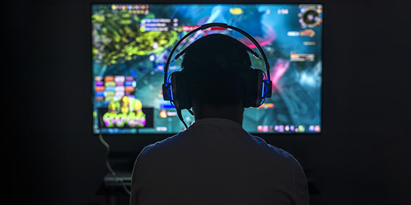 Silhouette of video gamer seen from behind, wearing headset. In front of the gamer is a brightly colored computer screen.