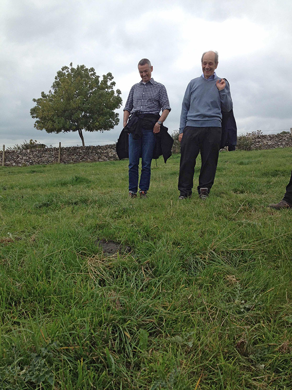 Two men stand in a green field looking down at a cow pat. A tree is in the background. It’s a cloudy day.