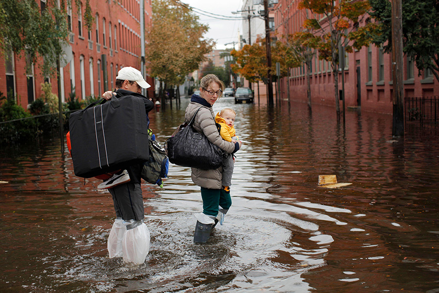 Photograph of a couple; the woman is holding a child. They are walking through a flooded street flanked by red brick buildings.