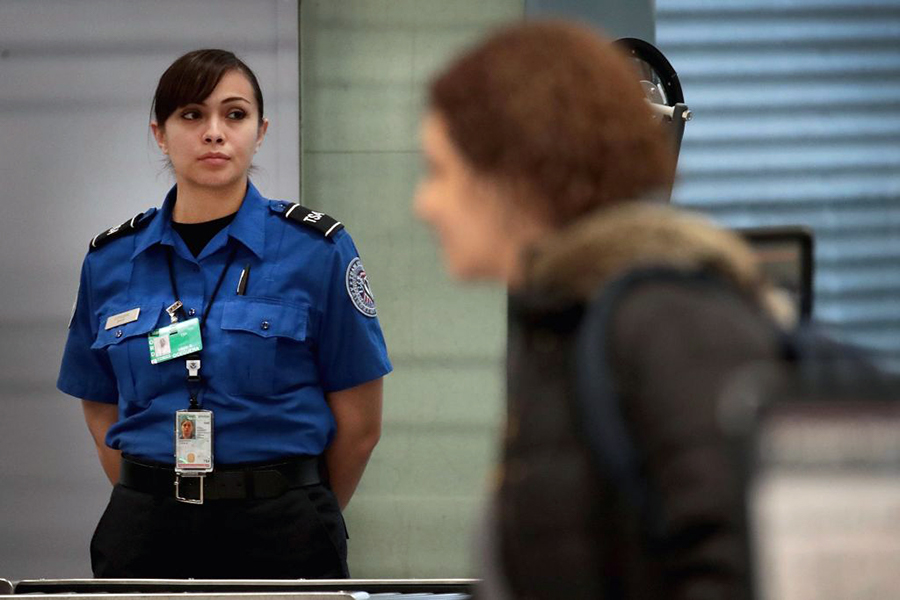 A security officer in uniform stands and watches a traveler, who is shown blurred in the foreground.