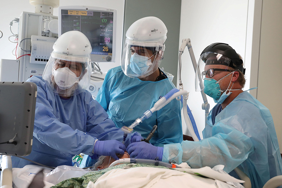 A photograph shows three healthcare workers dressed in masks, face shields, gowns and gloves around the bed of a patient in intensive care unit. They are intubating the patient and watching progress of the intubation on a screen.