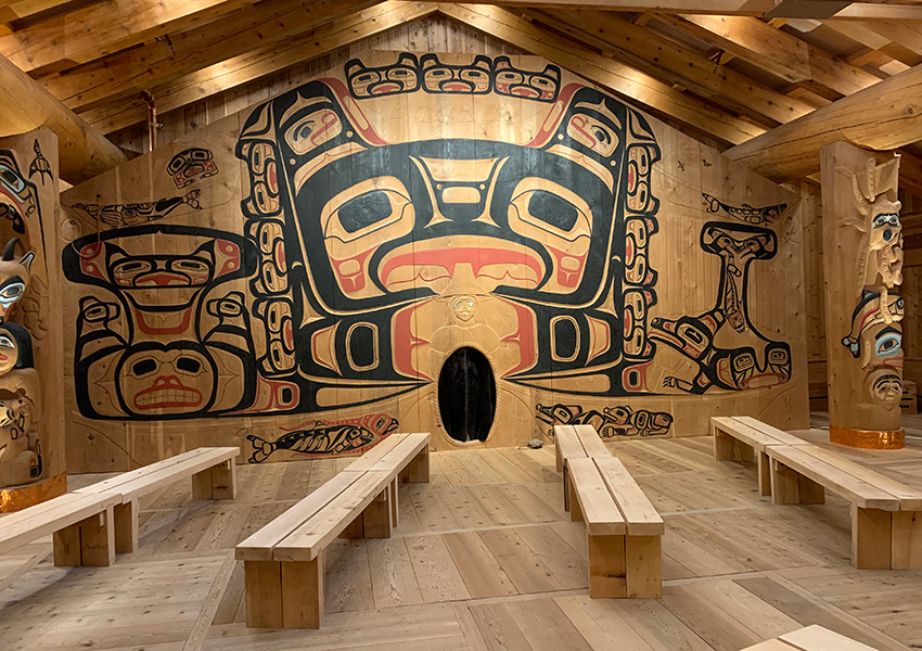 Photograph shows interior of a wooden hut with carvings, bright murals painted on the walls and a series of wooden benches.