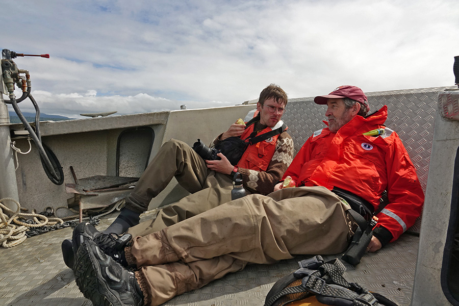 Two men lounge on the deck of a boat eating apples and talking. They are wearing hiking boots and warm clothing. The sky is cloudy.
