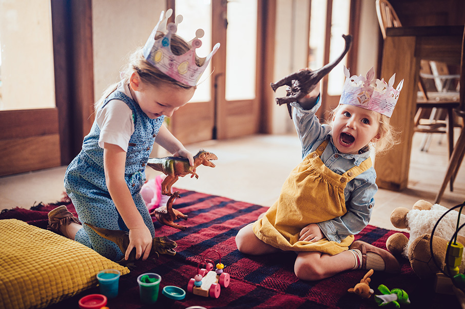 Photograph of two young children sitting on a blanket playing. Both are wearing paper crowns. One is yelling and waving a toy dinosaur in the air. The other is looking down, touching items on the blanket and also holding a dinosaur.