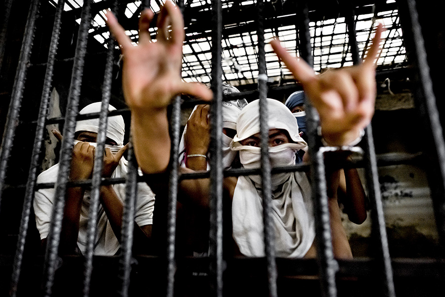 Young men inside a prison cage are shown with their faces covered with white T shirts. One of them, in the foreground, extends his arms through the metal bars, making hand signals.