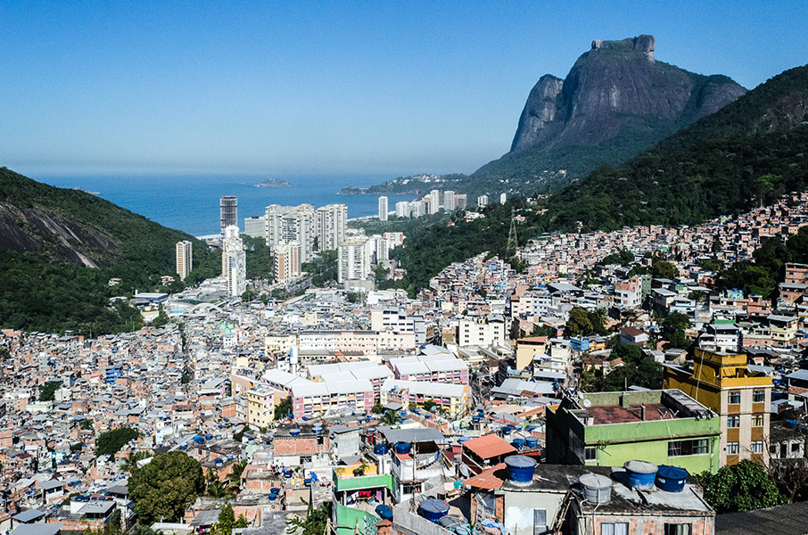 Aerial image of the Rocinha favela, in Rio de Janeiro, Brazil. Hundreds of multicolored, tightly spaced houses are tucked into a valley bordered by green mountains, with high-rise buildings and the sea visible in the distance. 