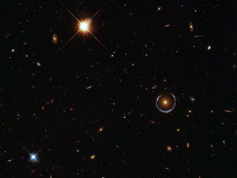 Hubble image shows a deep view of space with a horseshoe-shaped, bluish semi-circle around a bright glowing object visible among the stars and galaxies. Scientists believe the Horseshoe to be a galaxy with a redshift of about 2.4, meaning we see it as it was when the universe was just 3 billion years old.