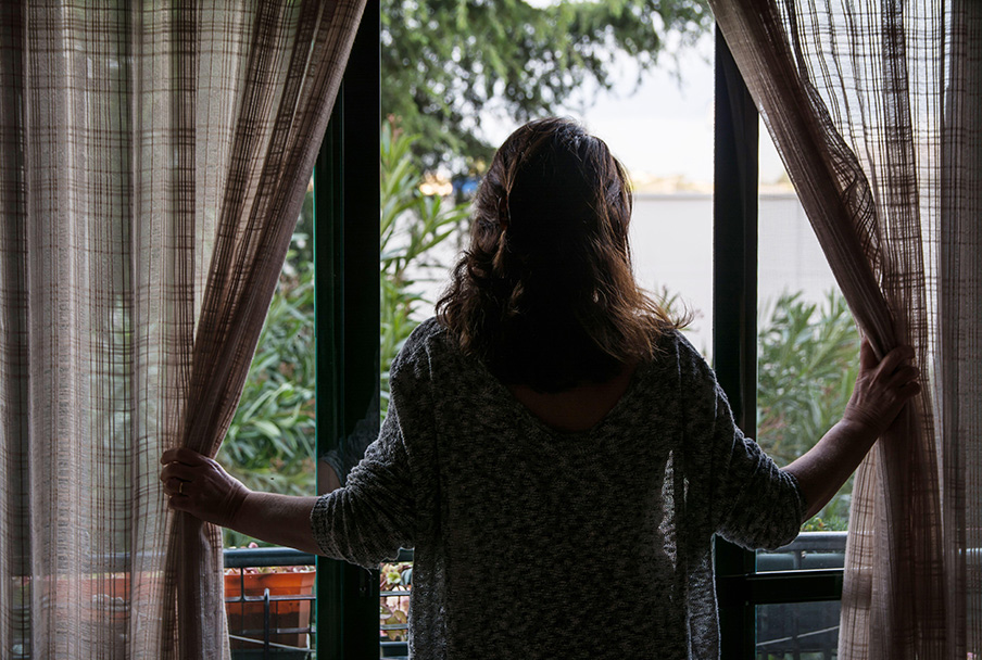 Photograph of a woman, from behind, standing in front of sliding doors of her balcony looking at the outdoors. She is holding curtains to one side. There is greenery beyond the balcony.