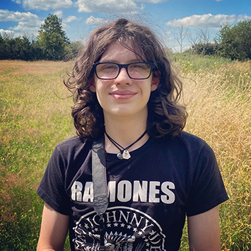 Photograph of a teenage boy standing in a field. He has brown, shoulder-length hair and is wearing glasses and a Ramones tee shirt.