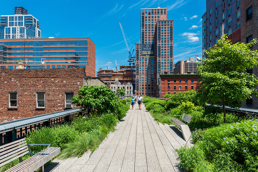 Photograph of two people walking along a broad elevated trail covered with gray flagstones. There are tall brick buildings on both sides of the trail, which is planted with small trees and other vegetation.