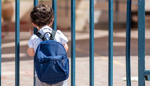 Fotografía de un niño pequeño con una mochila mirando a través de unas rejas el patio vacío de un colegio.