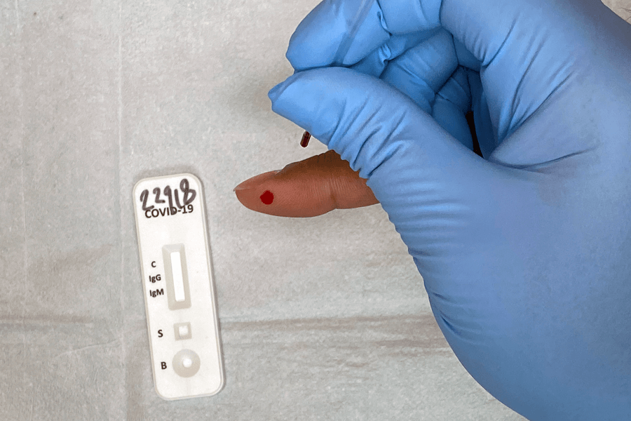 Close-up photograph of a hand wearing a blue glove. Underneath that person’s hand is the finger of an individual who is getting the test. The finger has been pricked and blood is being drawn into a small tube. To the left is the small testing strip to which the blood will be added.