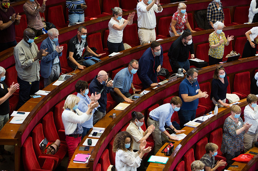 Casually dressed people wearing masks standing and applauding in a legislative chamber.