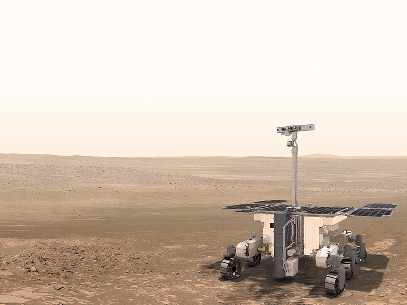 Photo of a cute, wheeled vehicle with an array of solar panels on its back. A projection that looks like a periscope is pointing upward. The feisty rover is trundling over a stark, dusty landscape.