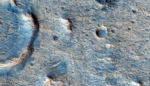 Blue-tinted image of a portion of the surface of Mars, with visible craters and pocks of different sizes.