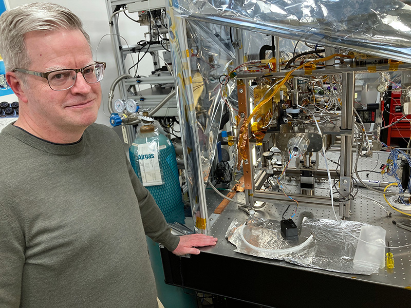 Photo of a man with glasses and olive-grey shirt standing next to a complicated-looking contraption with lots of wires, tubes and silvery reflective material.