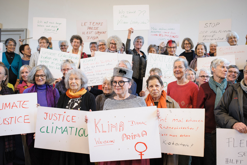 Photograph of a group of women, many with gray hair, standing together holding placards. The placards have phrases such as “Climate Justice” written on them.
