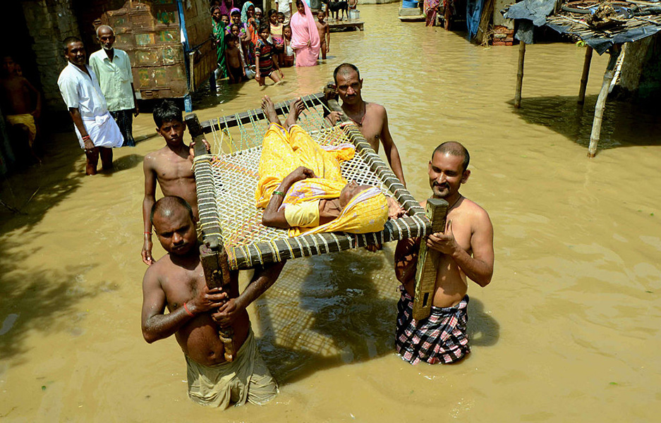 Photograph shows four men wading through muddy, thigh-high water. They are carrying a woman clad in yellow clothing on a stretcher. Men, women and children stand by in the water, watching.