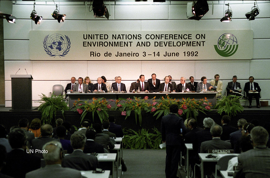 Photograph shows more than a dozen people sitting at a long, spot-lit table on a stage. A banner reads “United Nations Conference on Environment and Development, Rio de Janeiro 3-14 June 1992.” There are plants in front of the stage; the audience sit at desks.