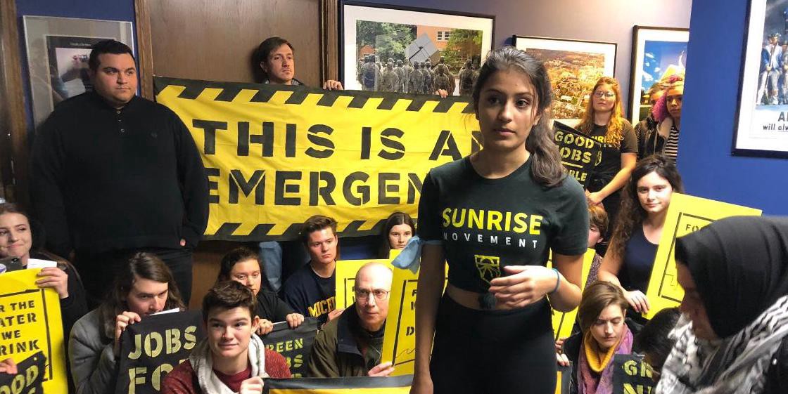 Photograph of young people in a hallway. One is holding a yellow sign that says, “This is an emergency.” A young woman stands at the front of the crowd wearing a black T-shirt with “Sunrise Movement” printed on it.