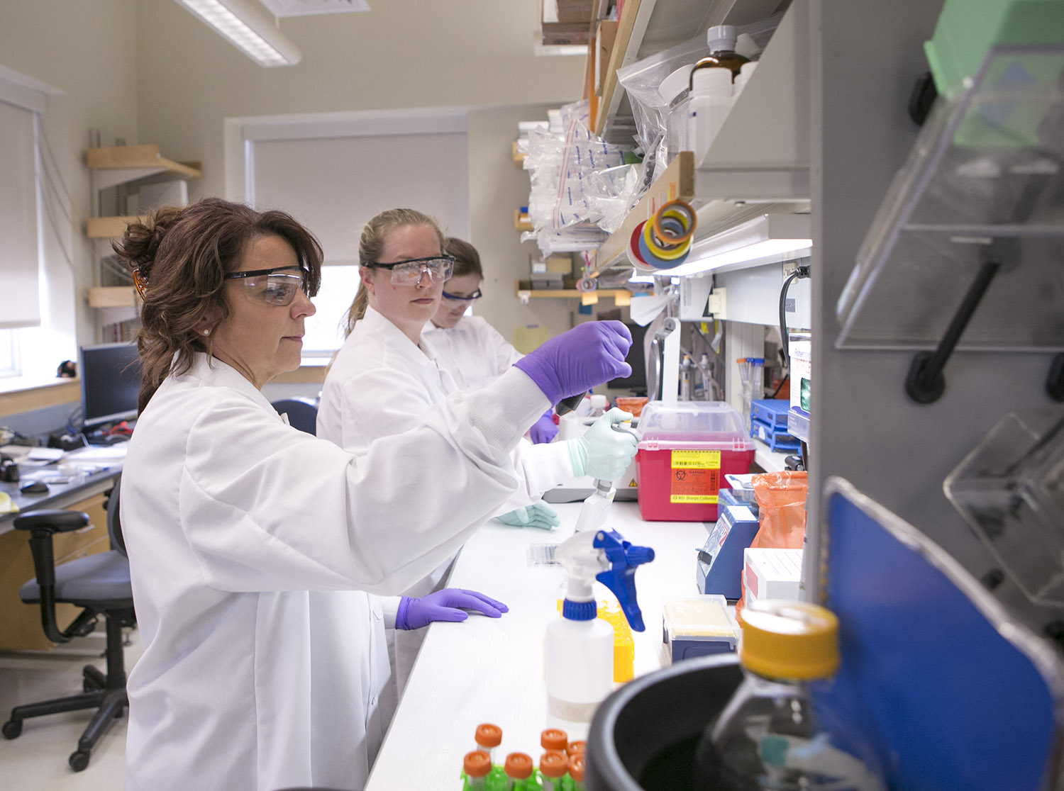 Photograph of three women wearing white coats, goggles and gloves and working at a lab bench.