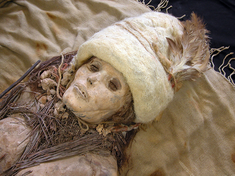 Close up photo of a mummy’s head and shoulders shows a skeleton wearing a hat with a feather, a string around its neck with small clumps tied to it, and twigs.