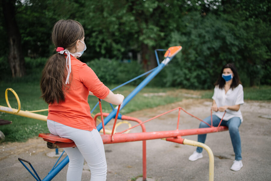 Photo shows a girl and a woman wearing protective masks playing on a see-saw at playground.