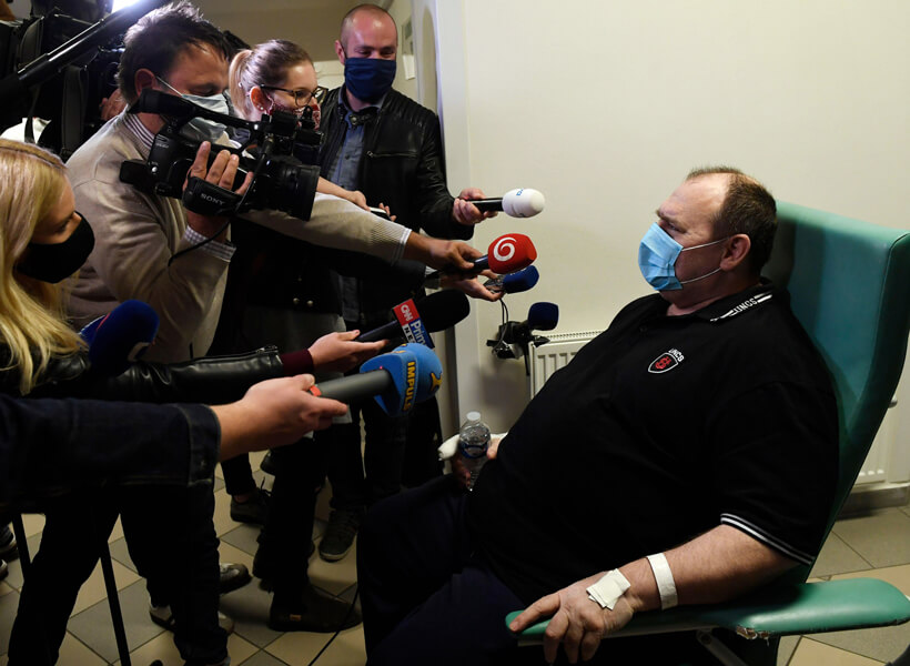 Photograph of a man sitting in a wheelchair wearing a surgical mask. To the left, a group of reporters seeking to interview him hold out microphones; one is holding a video camera.