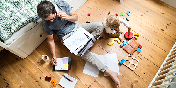 Photo shows a dad seated on the floor with a laptop balanced on his lap and writing in a planning calendar while talking on the phone. A baby sits at his knee, playing with blocks.