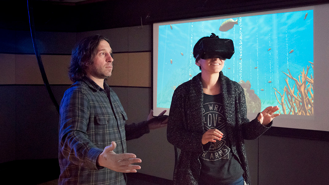 Photograph of a smiling woman wearing goggles over her eyes, her arms in front of her. A man stands to one side ready to catch her should she trip and fall. Behind them, a projector screen shows an underwater scene with fish and other marine life.
